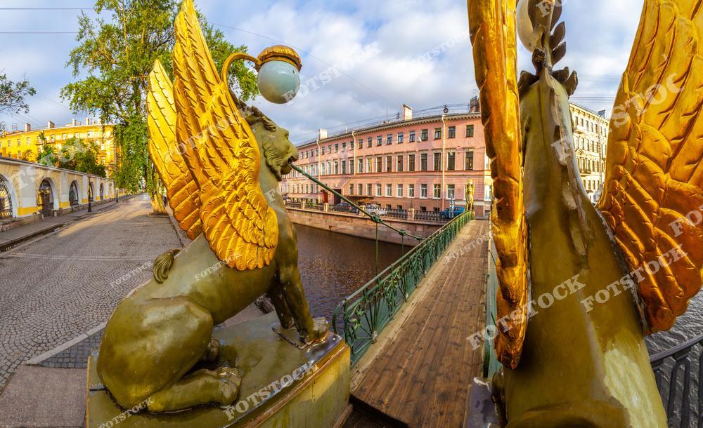 На фотографии изображены знаменитые грифоны с золотыми крыльями, украшающие Банковский мост через канал Грибоедова в Санкт-Петербурге. Этот небольшой пешеходный мост соединяет два берега канала и является одним из символов города. В светлое время суток золотые крылья грифонов особенно ярко сияют, подчеркивая архитектурное богатство исторического центра Петербурга. Вид на окружающие здания и мост создает атмосферу классического Петербурга.